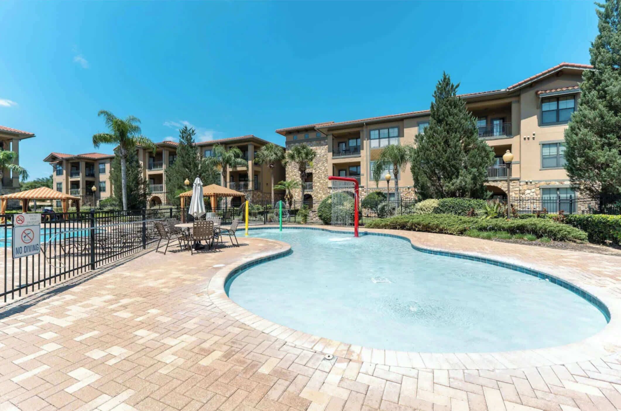 A swimming pool with a no diving sign, surrounded by lounge chairs and umbrellas, with multi-story buildings in the background.