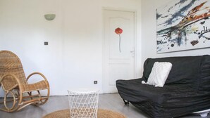 A room with a black sofa, a wicker chair, a small white table, and a door with a red flower decoration. - Villa cosy proche Valenciennes (La Sentinelle)