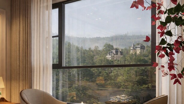 A room with a large window, a sofa, a small round table with teapots, and a view of a forested area with a building.