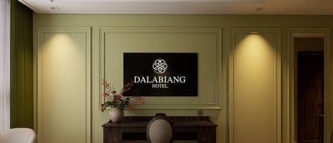 A hotel room with a bed, a desk with a chair, a potted plant, and a wall-mounted sign.