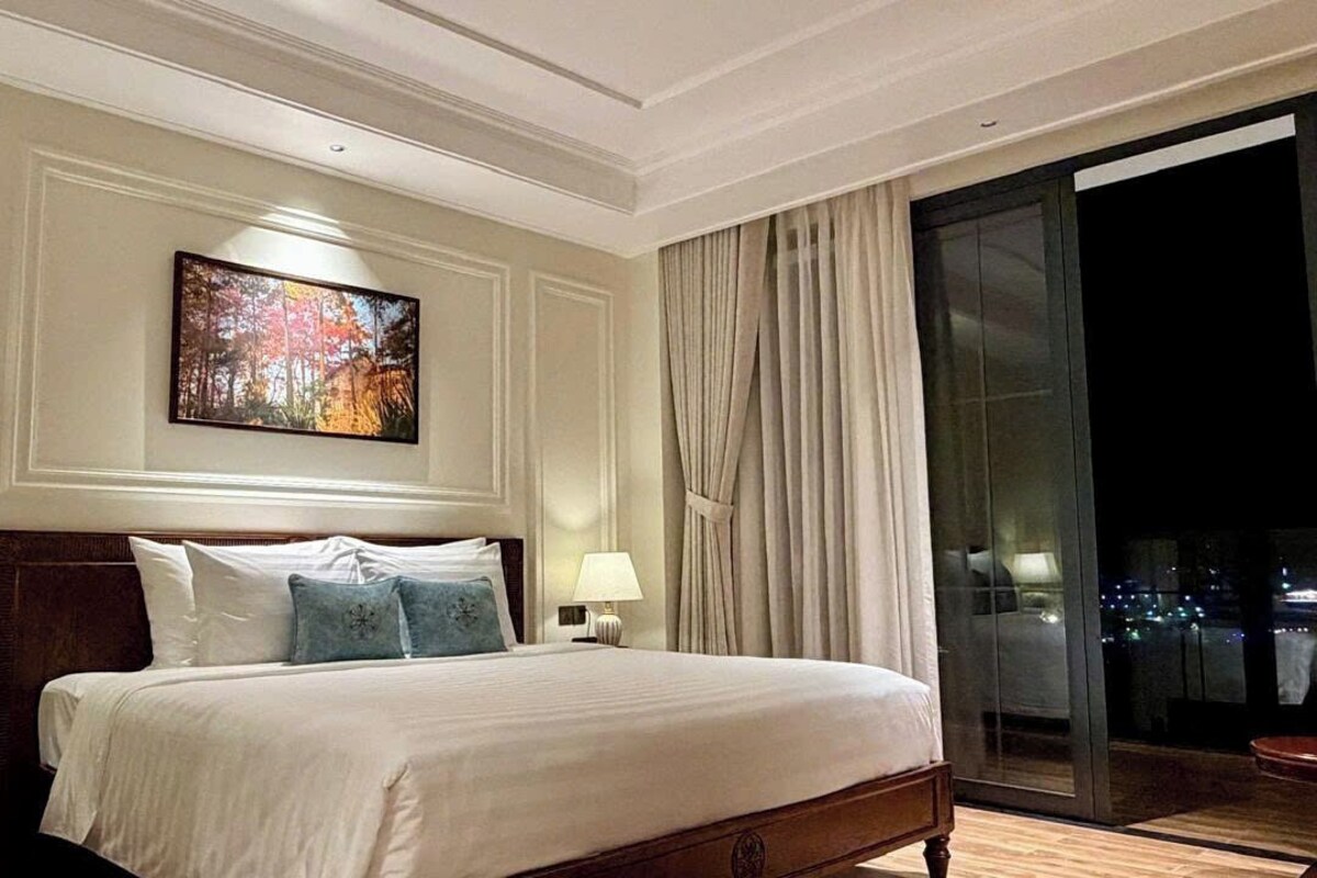 A hotel room with a large bed, two bedside tables with lamps, a painting above the bed, and a view of the city through a sliding glass door.