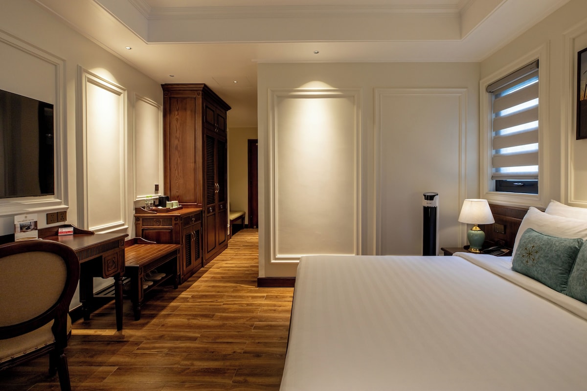 A hotel room with a large bed, a desk with a chair, a wardrobe, a bedside table with a lamp, and a window with blinds.
