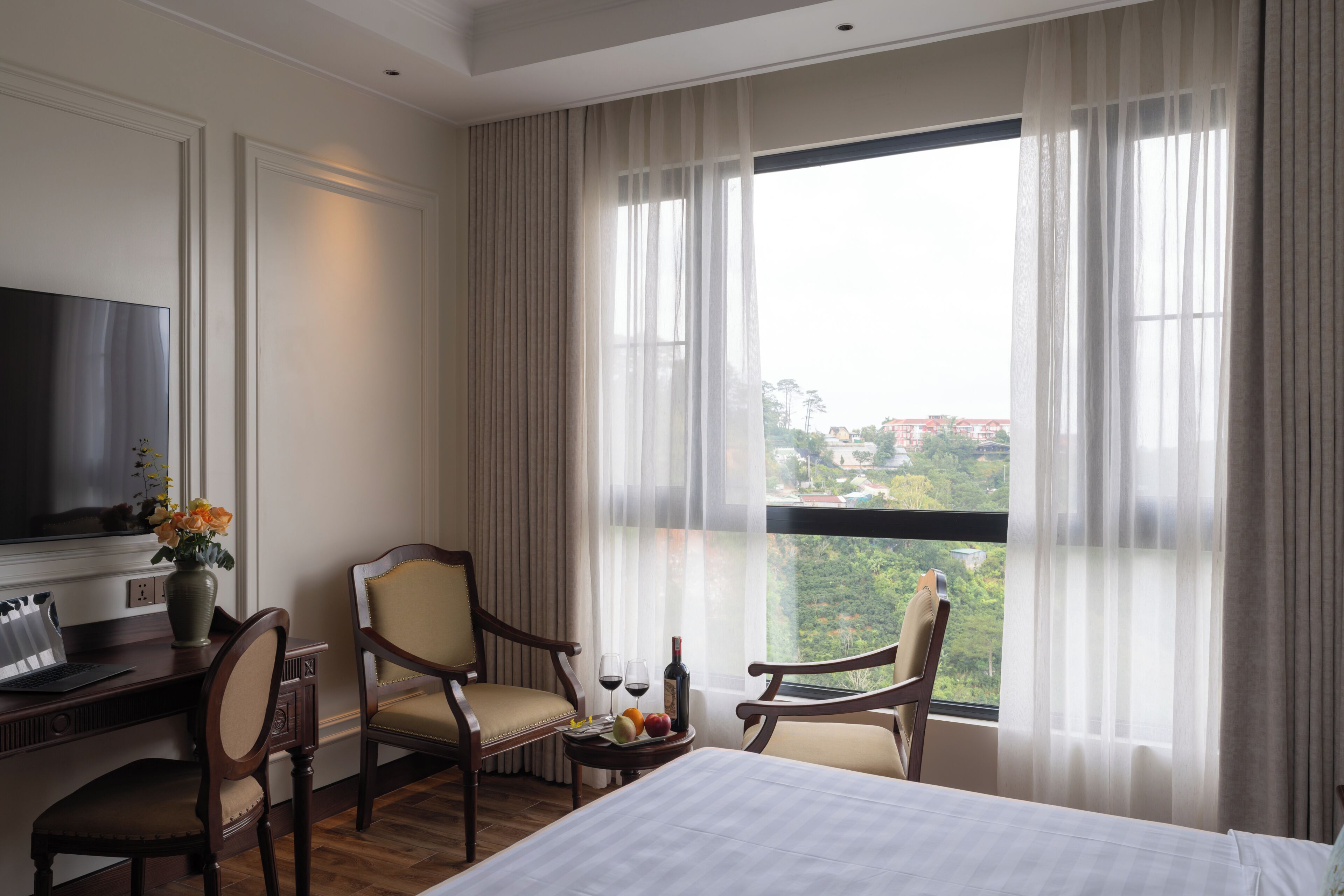 A hotel room with a bed, two armchairs, a desk with a laptop, a television, and a large window with curtains.