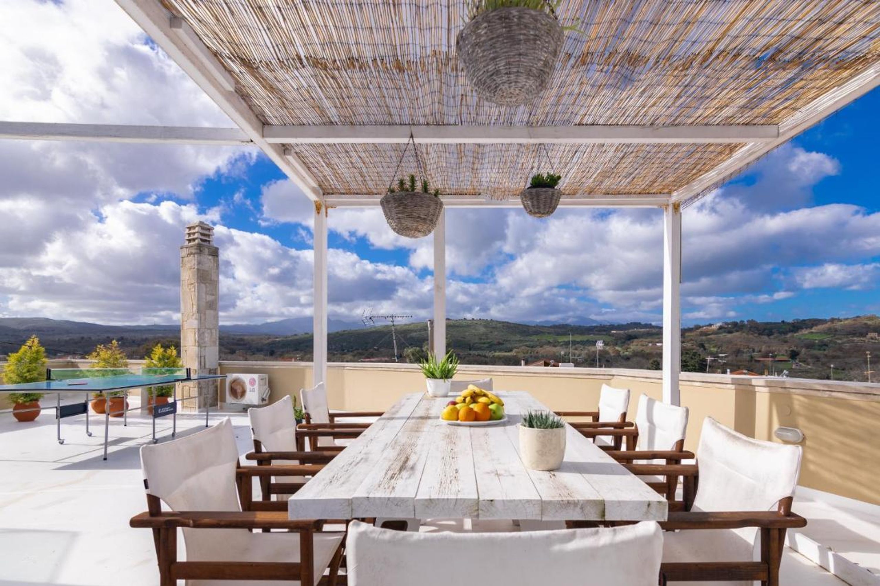 Una terraza en la azotea con una mesa de madera, sillas, cestas colgantes y vistas al paisaje.