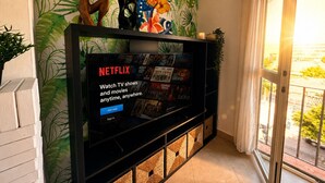 A living room with a TV displaying Netflix content, a wall with tropical leaf patterns, and a balcony with a view. - Stylish Flat in Puerto Banús - Steps to Marina (Marbella)