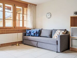 A room with a grey sofa, a clock on the wall, and a window with curtains.