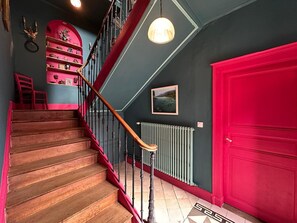 Eine Treppe mit Holzboden und roter Geländer, eine rote Tür, ein Heizkörper und ein an der Wand hängendes Bild.
