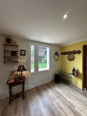 Een kamer met een houten vloer, een deur met ruitpanelen, een lamp op een tafel, een plank met spullen, en een muur met een krans en een hangend object.