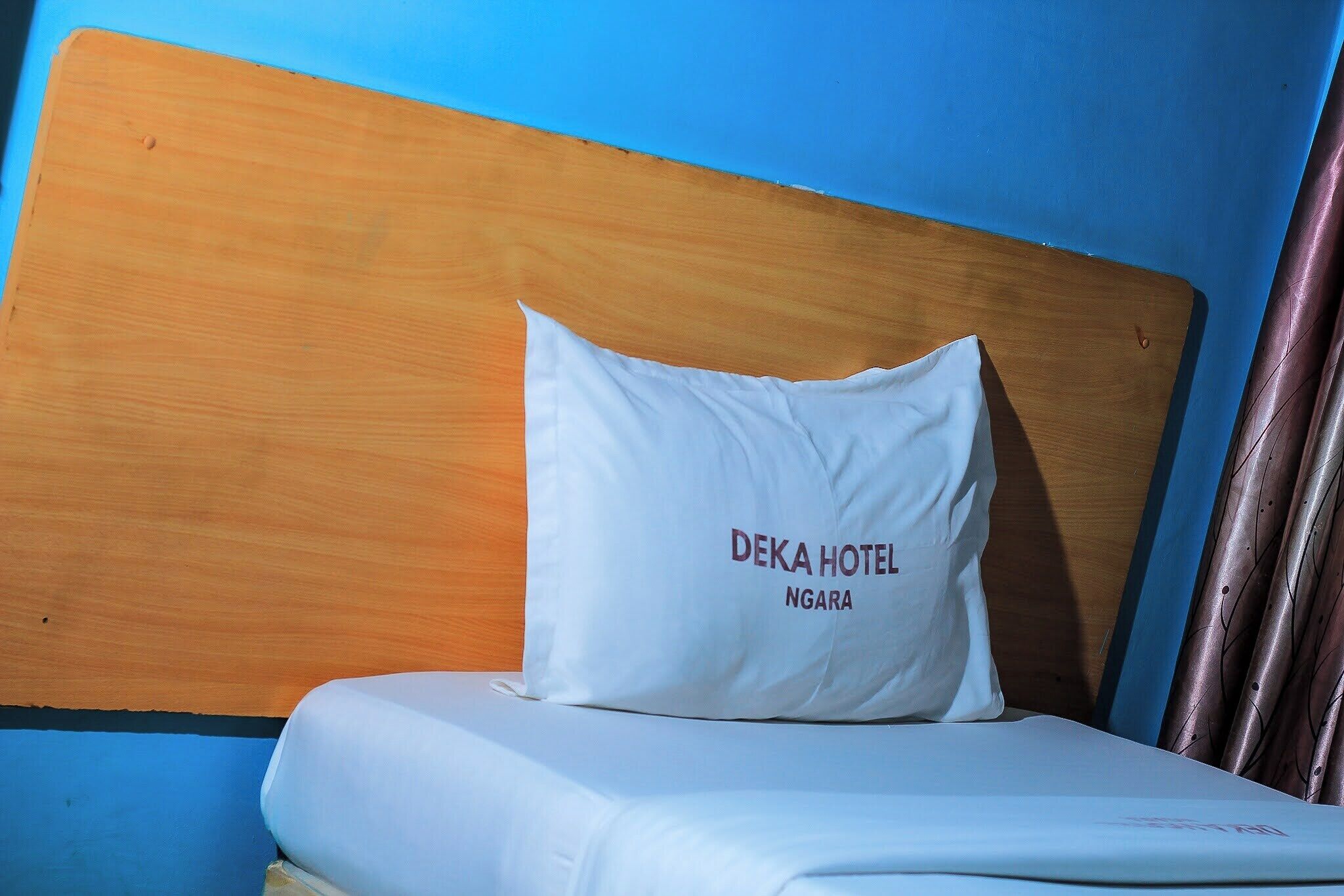 Ein Bett mit einem weißen Kopfkissen, beschriftet mit 'DEKA HOTEL NGARA', und einem hölzernen Kopfteil.
