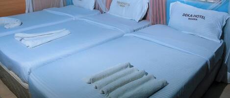 Quarto de hotel com duas camas, cada uma com uma almofada rotulada 'DEKA HOTEL NGARA', uma toalha dobrada sobre cada cama e uma janela com cortinas.