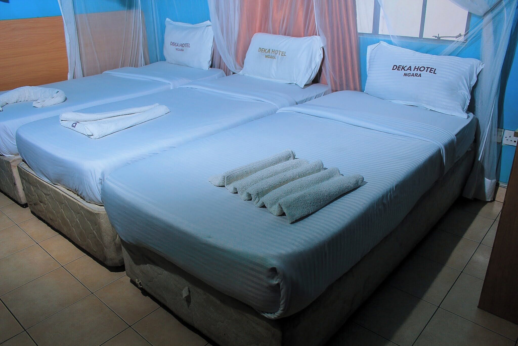 Habitación de hotel con dos camas, cada una con una almohada que dice 'DEKA HOTEL NGARA', una toalla doblada prolijamente en cada cama y un mosquitero cubriendo las ventanas.