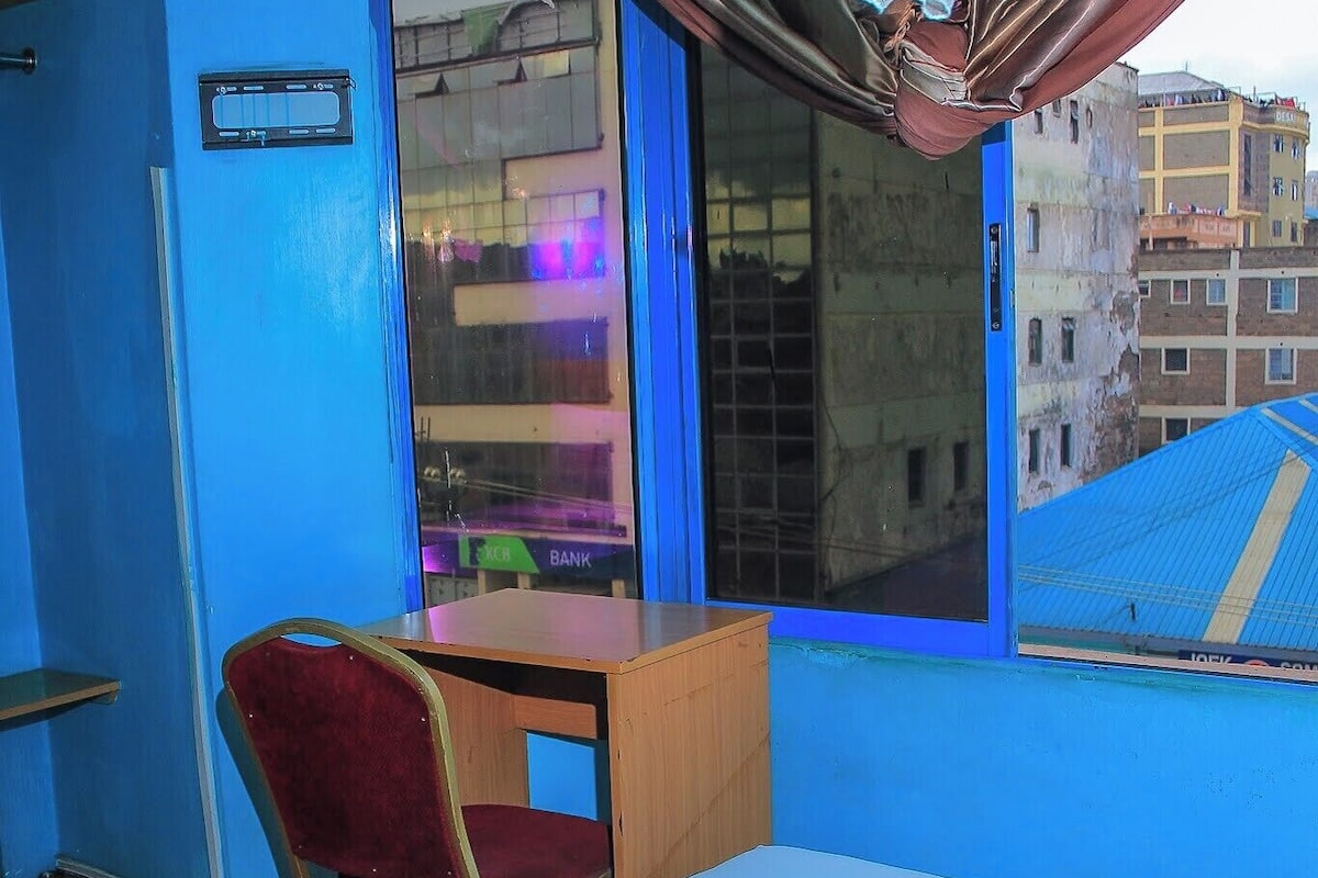 A hotel room with a bed, a desk, a chair, a window with curtains, and a view of buildings outside.