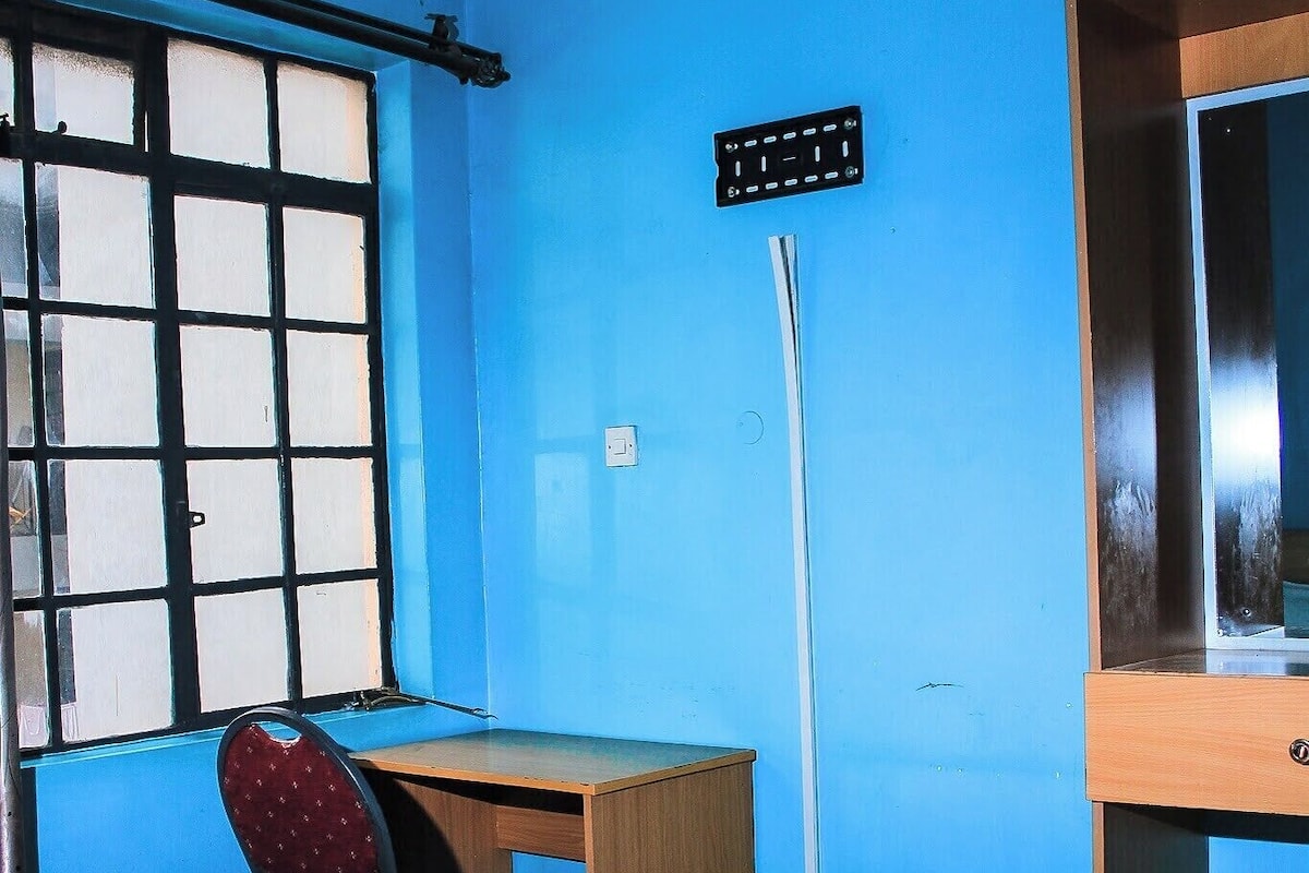 A room with a bed, a desk, a chair, a window with bars, and a wall-mounted control panel.
