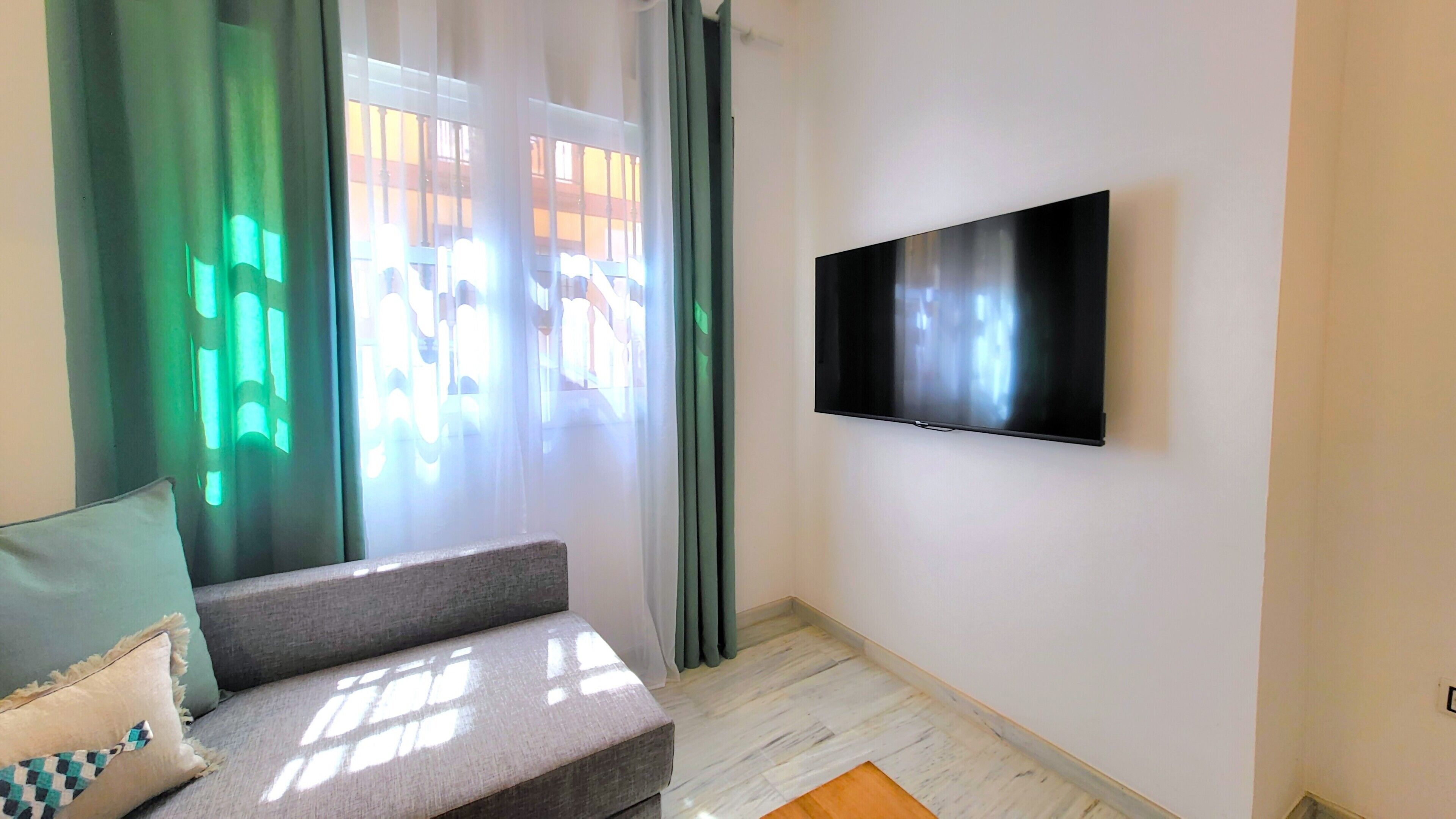 Un salon moderne avec un téléviseur à écran plat fixé au mur, un canapé gris agrémenté de coussins et une fenêtre ornée de rideaux verts.