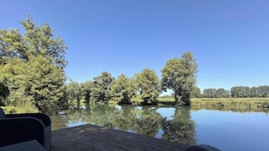 Ein ruhiger See mit Bäumen, deren Spiegelbild sich im Wasser zeigt, ein hölzerner Steg und ein klarer, blauer Himmel.