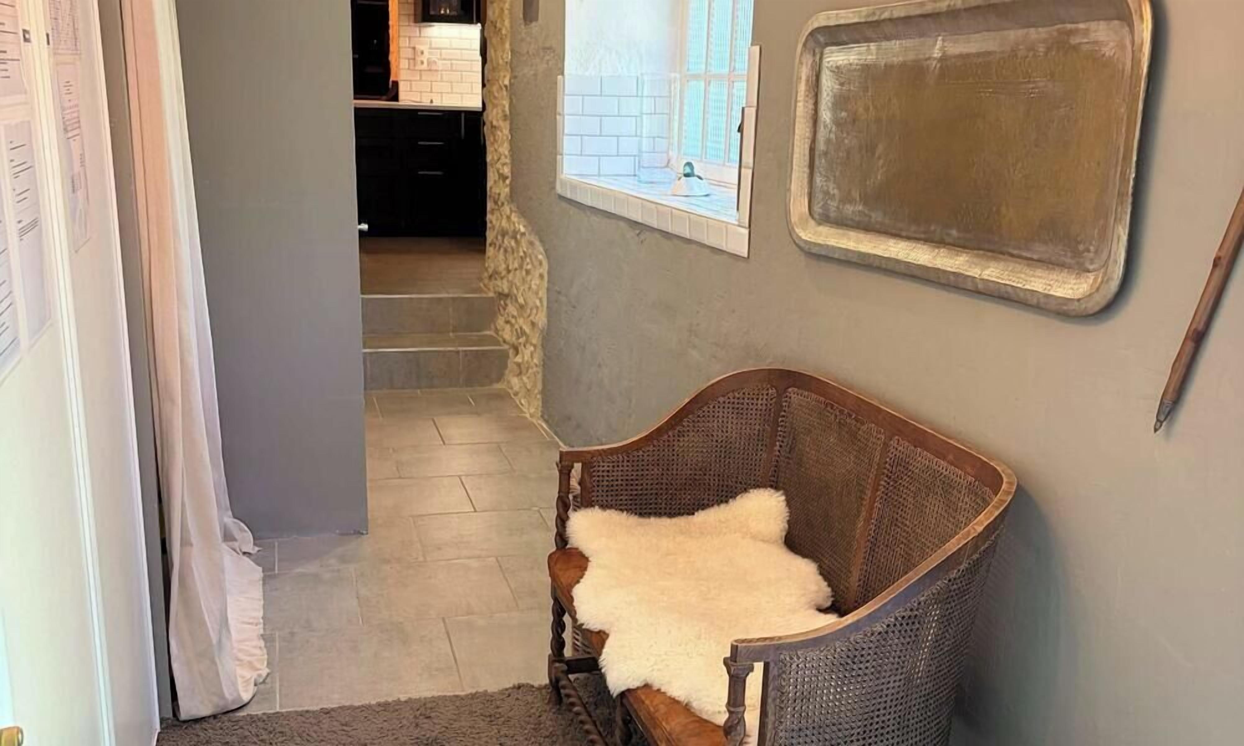 Una habitación con una silla de mimbre, un plaid blanco y mullido, una ventana con vistas y un espejo montado en la pared.