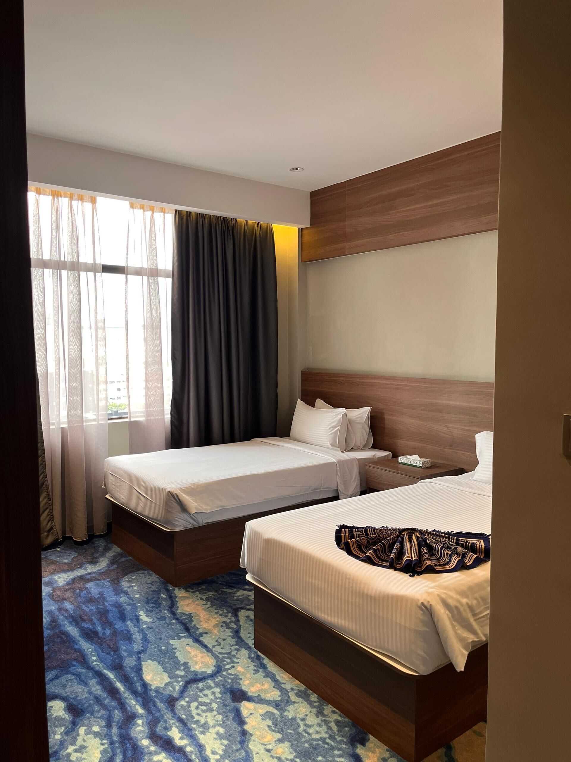 A hotel room with two beds, a large window with curtains, a wooden headboard, and a patterned carpet.