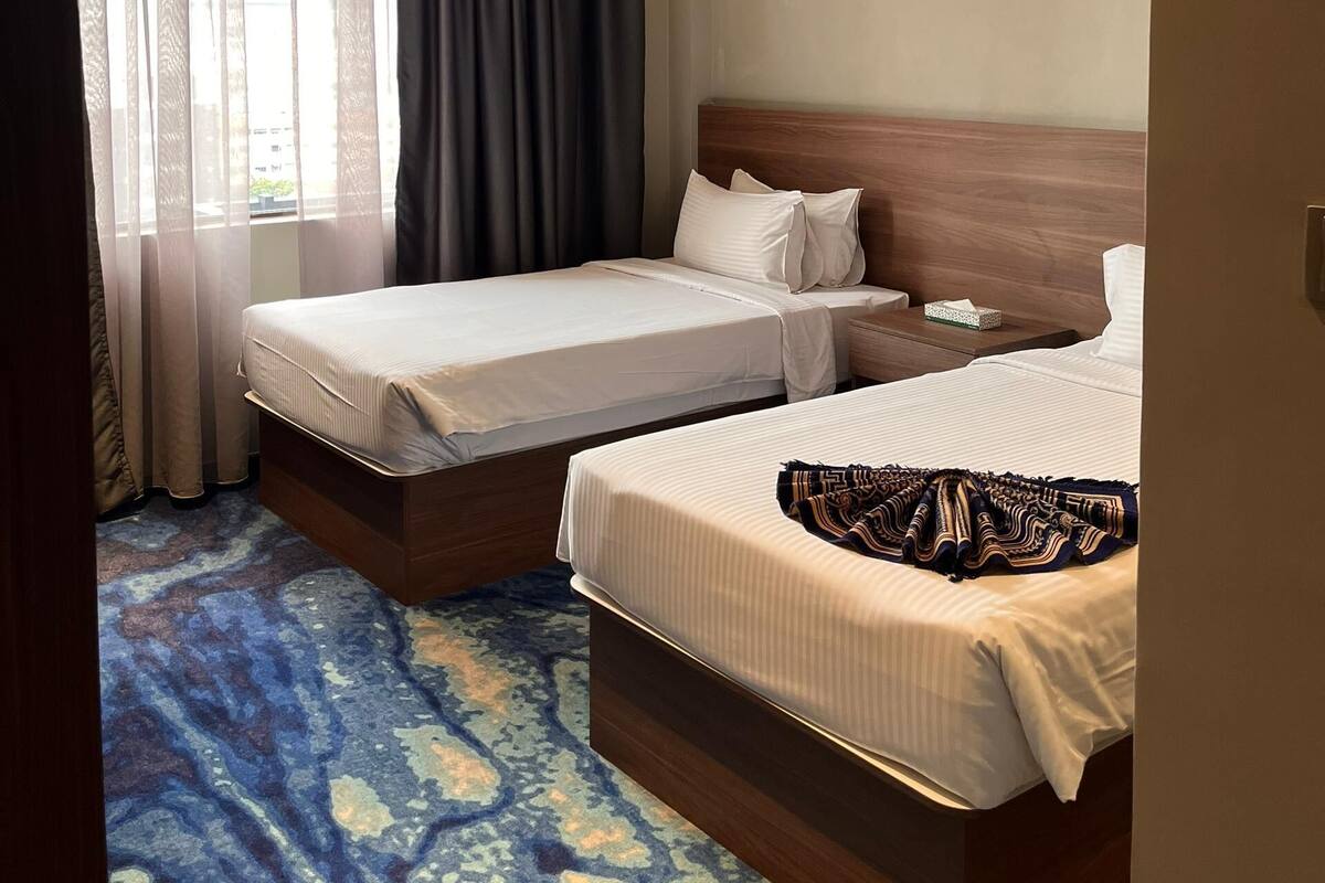 A hotel room with two beds, a large window with curtains, a wooden headboard, and a patterned carpet.