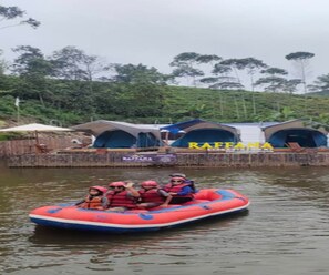 A raft with people wearing life jackets on a body of water, with tents labeled 'RAFFANA' in the background. - Cozzy Raffana Glamping (bandung)