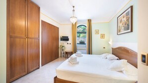 Une chambre avec un grand lit, une armoire en bois, un lustre, une télévision et une vue sur un jardin par la fenêtre.