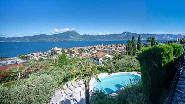 Une piscine entourée de verdure, avec vue sur une ville côtière et des montagnes à l’horizon.