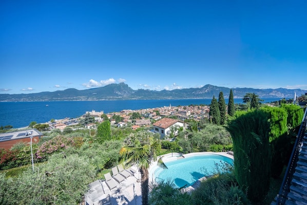 A pool surrounded by greenery, a view of a coastal town, and distant mountains. - ApartmentsGarda - Villa Giada - TOPAZIO (TORRI DEL BENACO)