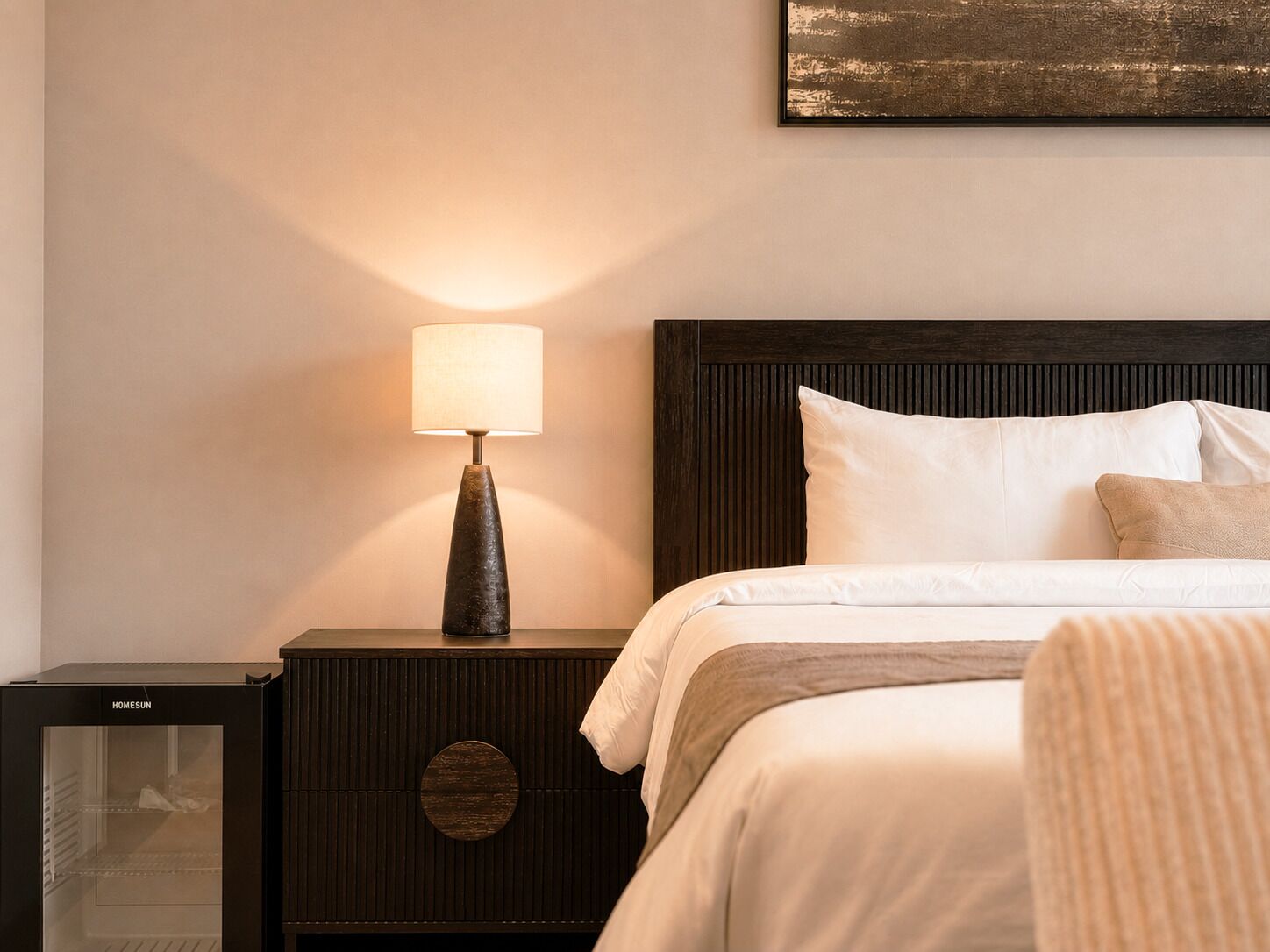 Una habitación de hotel con una cama, una mesita de noche con lámpara y un refrigerador.