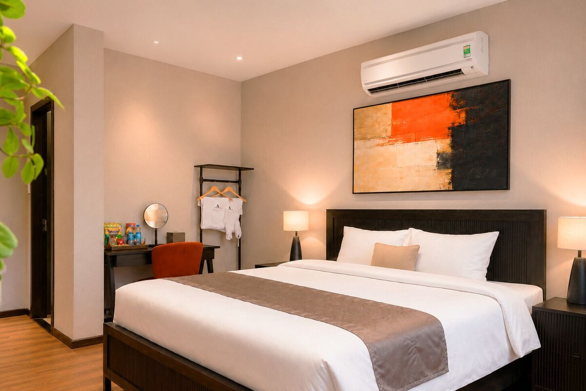 A hotel room with a large bed, bedside tables, a desk with a chair, a hanging towel rack, and an air conditioning unit.
