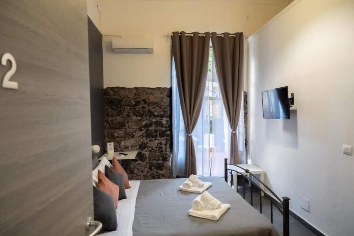 Una habitación de hotel con cama, televisor, pared de piedra y ventana con cortinas.