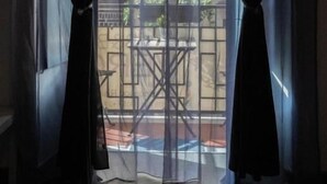 Una ventana con cortinas traslúcidas, vista a un espacio exterior con una mesa y sillas, y un edificio al fondo.