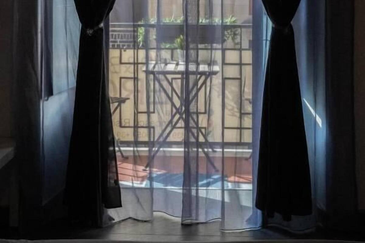 Una ventana con cortinas traslúcidas, vista a un espacio exterior con una mesa y sillas, y un edificio al fondo.
