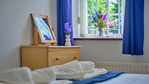 Ein Schlafzimmer mit einem Bett, einem Nachttisch mit einem gerahmten Bild und einer Blumenvase sowie einem Fenster mit Vorhängen.