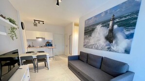 Una sala de estar moderna con un sofá gris, una zona de comedor con sillas, una cocina con armarios blancos y un cuadro grande de un faro en la pared.