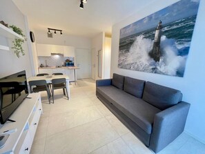 A modern living room with a grey sofa, a dining area with chairs, a kitchen with white cabinets, and a large painting of a lighthouse on the wall. - Sea and pool view holiday stay (Porto Santa Margherita)