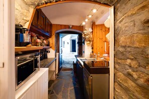 A narrow kitchen with wooden cabinets, a stone wall, and a view into a dining area. - [Courmayeur City Center] Imperial Elegance (Courmayeur)
