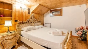 Quarto com teto de madeira, cama com cabeceira decorada, mesa de cabeceira com abajur e radiador.