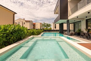 En swimmingpool med flisebelagt område, omgivet af grønt og moderne bygninger.