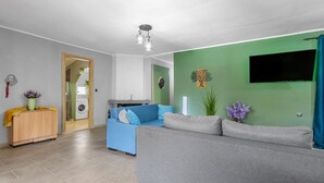 A living room with a grey sofa, a blue sofa, a wooden cabinet, a TV, and a green wall. - Elena s House in Mournies (Chania)