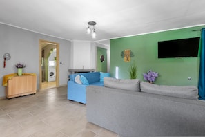 A living room with a grey sofa, a blue sofa, a wooden cabinet, a TV, and a green wall. - Elena s House in Mournies (Chania)