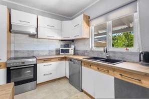 A modern kitchen with white cabinets, a stainless steel oven, a dishwasher, a microwave, and a window with a view of greenery. - Elena s House in Mournies (Chania)