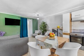 A modern living room with a flat-screen TV, a dining table with chairs, a sofa, a potted plant, and a kitchen area with a stove. - Elena s House in Mournies (Chania)