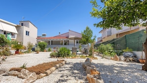 A residential area with a house featuring a red-tiled roof, a covered patio, and a garden with various plants and a gravel driveway. - Elena s House in Mournies (Chania)