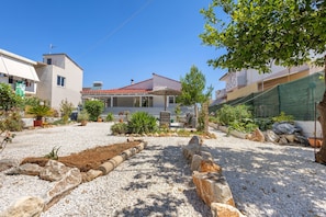A residential area with a house featuring a red-tiled roof, a covered patio, and a garden with various plants and a gravel driveway. - Elena s House in Mournies (Chania)