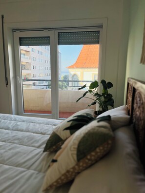A bedroom with a bed, a window with a view of a building, and a plant on the windowsill. - Casa Mar - Beautiful apartment with an ocean view on Matosinhos Beach in Porto (Matosinhos)
