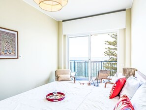 Hotellihuone, jossa on sänky, pieni pöytä lasineen, tuoli, sohva, parveke kaiteineen ja taulu seinällä.