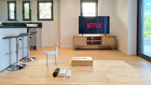 Una sala moderna con área de cocina, un televisor grande mostrando 'NETFLIX' y una mesa de centro de madera con controles remotos y una caja de pañuelos.
