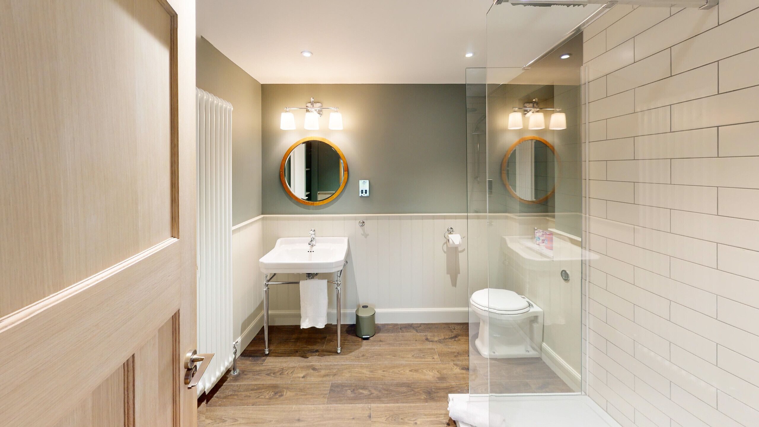 A bathroom with a wooden floor, a white sink, a toilet, and a glass-enclosed shower area.