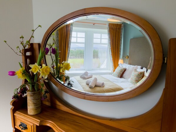 A bedroom with a large mirror, a bed with towels, a vase with flowers, and a view of the outdoors.