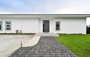 A modern building with a flat roof, a central entrance with a dark door, and a paved driveway leading to the entrance. - Konmek Poolvilla (Sattahip)