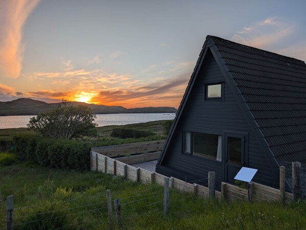 A modern house with a dark roof, large windows, and a view of a lake at sunset.